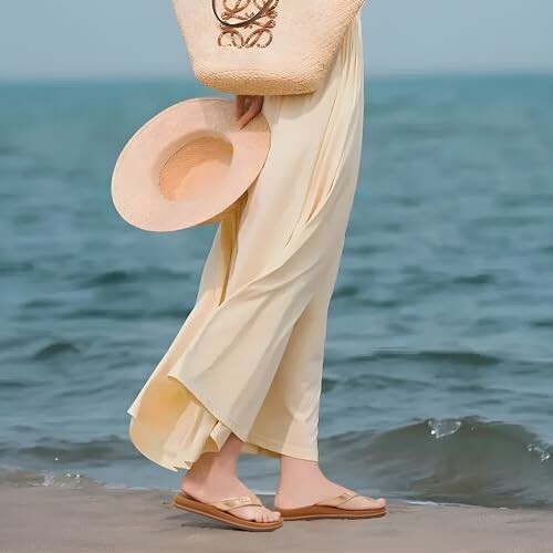 Person in a flowing dress holding a sun hat near the ocean